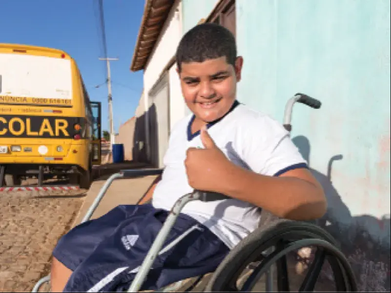Menino, Niltomar, sentado em uma cadeira de roda faz sinal de legal ao fundo ônibus escolar