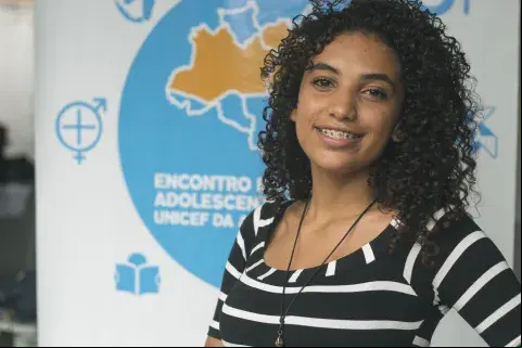 Jovem olha para frente e sorri. Ela veste blusa preta com listras brancas, cabelo solto e usa aparelho nos dentes. Ao fundo, um  branco com detalhes em azul indica que ali é um encontro de adolescentes do NUCA